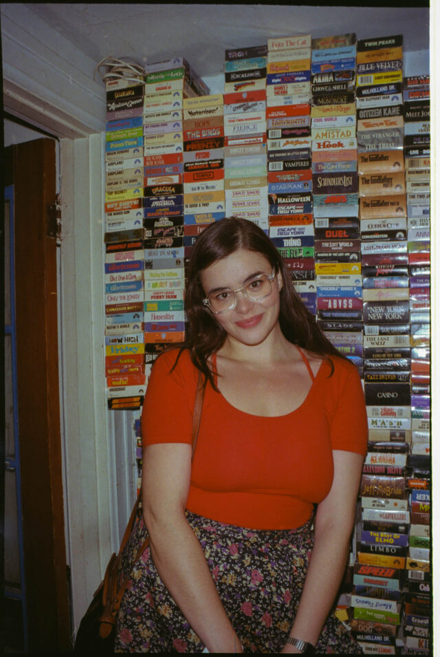 A scene from Mile End Kicks. Grace (Barbie Ferreira) stands in front of a wall of VHS tapes.