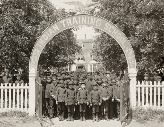A group of boys stand under an arch for an Indian Training School.