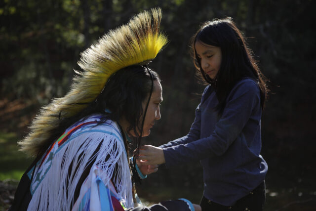 A Native American man kneels as his daughter fastens a headdress on him.