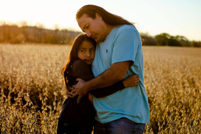 A Native American man hugs his daughter in a field.